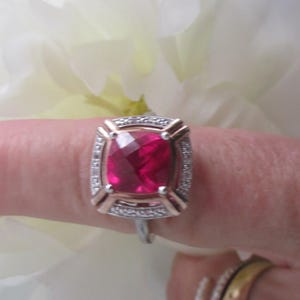 May include: A ring with a large, red, cushion-cut gemstone surrounded by a halo of smaller, white gemstones. The ring is set in a gold band.