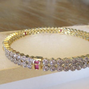 May include: A gold bangle bracelet featuring two parallel rows of round clear stones and evenly spaced rectangular red stones set in a metallic frame. The design consists of a continuous circular band with a repeating pattern of stones.