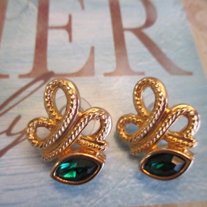 May include: A pair of gold tone earrings with a green gem stone in the center of each earring. The earrings are shaped like a bow with a rope-like design.