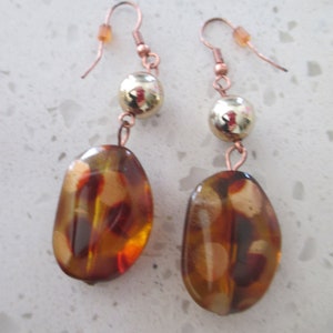 May include: A pair of dangle earrings with copper wire and brown, orange, and gold glass beads. The beads are oval shaped and have a mottled pattern.