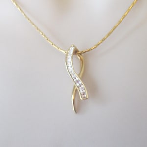 May include: A gold-toned necklace with a pendant featuring a curved design adorned with small, clear gemstones. The chain is delicate and also gold-toned. The jewelry is displayed against a soft, neutral background.