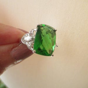 May include: A silver ring with a large, emerald green gemstone. The ring has a delicate, ornate design.