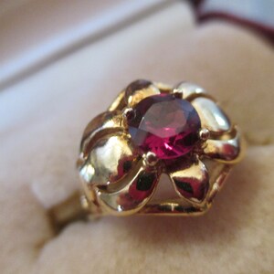 May include: A gold-colored ring with a flower-shaped setting. The ring features a round, deep red gemstone at its center. The setting has curved petals, and the ring is set against a neutral background.