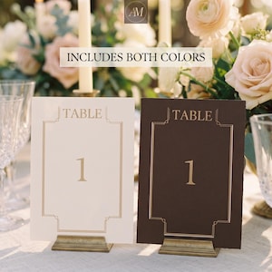 May include: Two rectangular table number cards, one cream and one dark brown, each featuring the text TABLE 1 in gold foil with a matching gold geometric border. Both cards are held upright by small, rectangular, metallic gold stands.