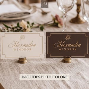 Puede incluir: Dos tarjetas de lugar rectangulares sobre soportes dorados. Una tarjeta es crema con texto oscuro, la otra es marrón oscuro con texto dorado. Ambas muestran una ilustración floral, el nombre Alexandra Windsor y el texto TABLE 01.