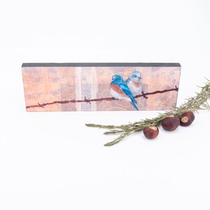May include: A rectangular wooden sign with a painted image of two bluebirds perched on a barbed wire fence. The background is a textured, peach-colored design.