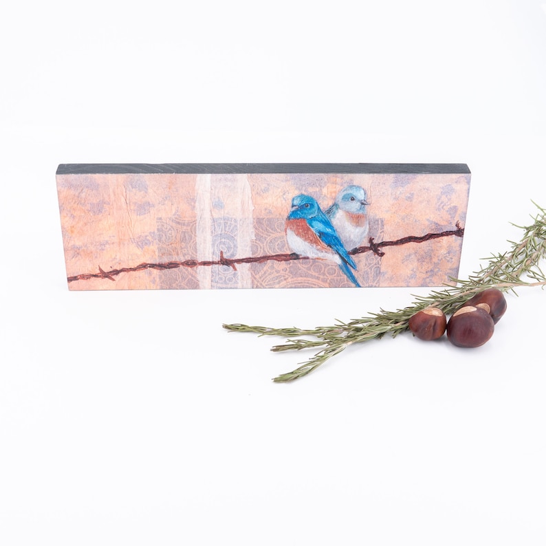 May include: A wooden plaque with a painted image of two bluebirds perched on a barbed wire fence. The background is a soft peach color with a white and brown patterned design.