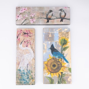 May include: Set of four rectangular wood wall art panels with a nature theme. The first panel features a white bird with pink flowers. The second panel features a blue bird perched on a sunflower. The third panel features two brown birds on a branch with pink flowers. The fourth panel features a blue bird perched on a sunflower.