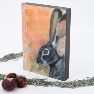 May include: A black-tailed jackrabbit with large ears is painted on a wood panel with a textured orange background. The painting is framed with a dark wood frame.