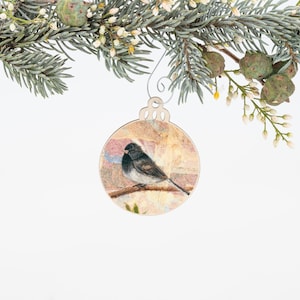 May include: Wooden Christmas ornament featuring a watercolor-style image of a small bird perched on a branch. The ornament has a round shape with a decorative metal hanger and is set against a backdrop of evergreen branches and white background.