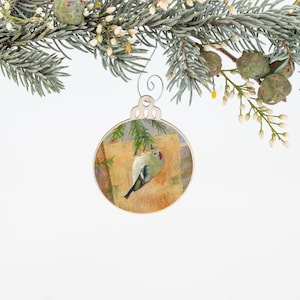 May include: A round wooden Christmas ornament with a painted image of a small bird perched on a branch. The bird is green and white with a red spot on its head. The background is a light brown with a hint of green.