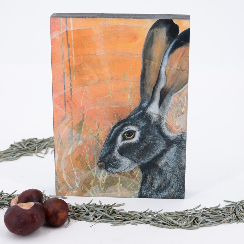 May include: A close-up of a painting of a black-tailed jackrabbit with a brown and orange background. The rabbit's fur is a dark gray with white markings around its eyes. The background is a mix of orange, brown, and white.