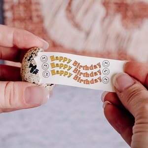 May include: A white paper strip with the text "Happy Birthday" repeated three times, with a small smiley face icon next to each phrase. The strip is being peeled off of a small, speckled, brown quail egg.