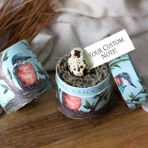 May include: A decorative gift box set with a bird theme. The set includes a cylindrical box with a bird and nest design, a matching lid, and a small card that reads 'Your Custom Note!'. Inside the box is a nest of hay with a speckled egg. The box is light blue with a bird illustration.