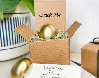 May include: A gold-painted egg in a brown cardboard box with the words "Crack Me" on the lid. The egg is surrounded by shredded paper. A card with the text "Thank You Mom For Everything" is next to the box. The egg is broken into pieces.