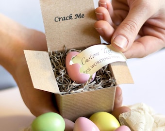 May include: A small, pink, ceramic egg with a crack in it is nestled in a brown box with the words "Crack Me" printed on the lid. The box is surrounded by a variety of pastel-colored eggs.