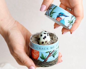 May include: A round light blue cardboard container with a lid, decorated with illustrations of birds and branches. The text CRACK ME is printed on the side. Inside, a speckled quail egg rests on a bed of dried grass fibers, partially revealed.