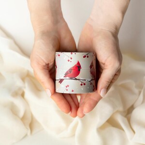 Puede incluir: Una caja de regalo cil&iacute;ndrica con un p&aacute;jaro cardenal rojo posado en una rama nevada con bayas rojas. La caja se sostiene con dos manos sobre un fondo blanco roto. La caja tiene aproximadamente 5 cm de di&aacute;metro.