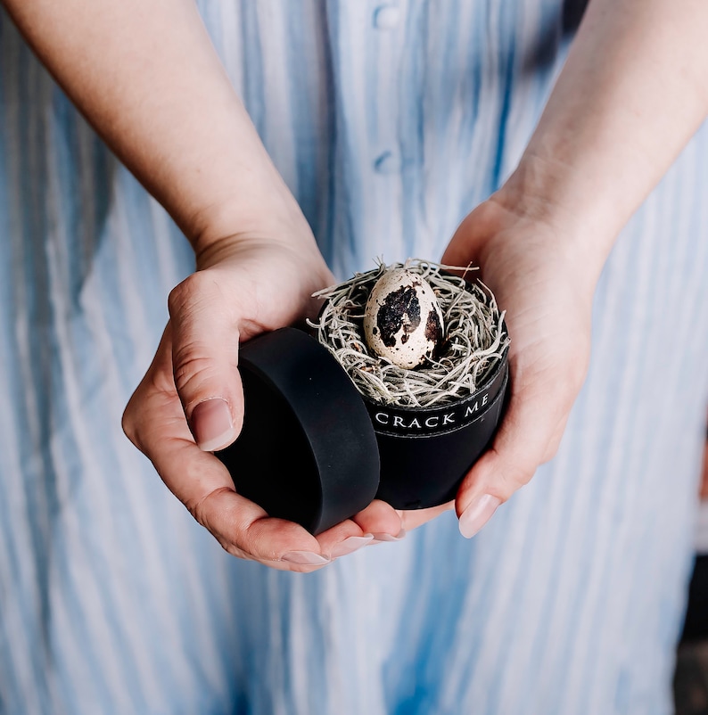 May include: A small, round black box with the text "CRACK ME" holds a speckled quail egg nestled in a bed of dried grass. The box is open, revealing the egg. The image is taken from a close-up angle.