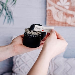 Pu&ograve; includere: Una scatola regalo rotonda nera con la scritta "CRACK ME" stampata sul lato. La scatola contiene un oggetto nero e un'imbottitura decorativa. Viene tenuto in mano un piccolo biglietto con "Happy Anniversary!" e un cuore rosso.