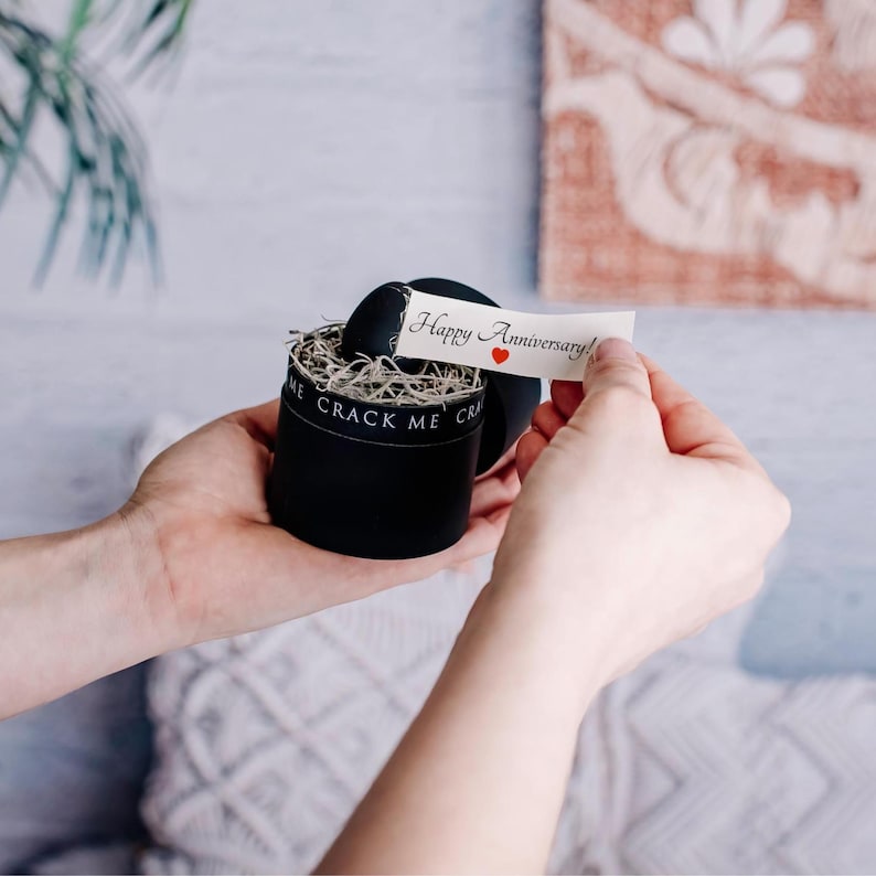 Pu&ograve; includere: Una scatola regalo rotonda nera con la scritta "CRACK ME" stampata sul lato. La scatola contiene un oggetto nero e un'imbottitura decorativa. Viene tenuto in mano un piccolo biglietto con "Happy Anniversary!" e un cuore rosso.