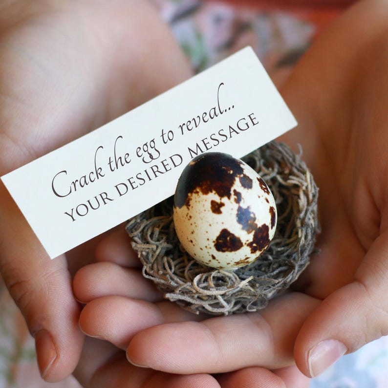 May include: A speckled egg rests in a small nest, held in cupped hands. A white card above the egg reads "Crack the egg to reveal... YOUR DESIRED MESSAGE". The egg has brown spots and is surrounded by a nest made of twigs.