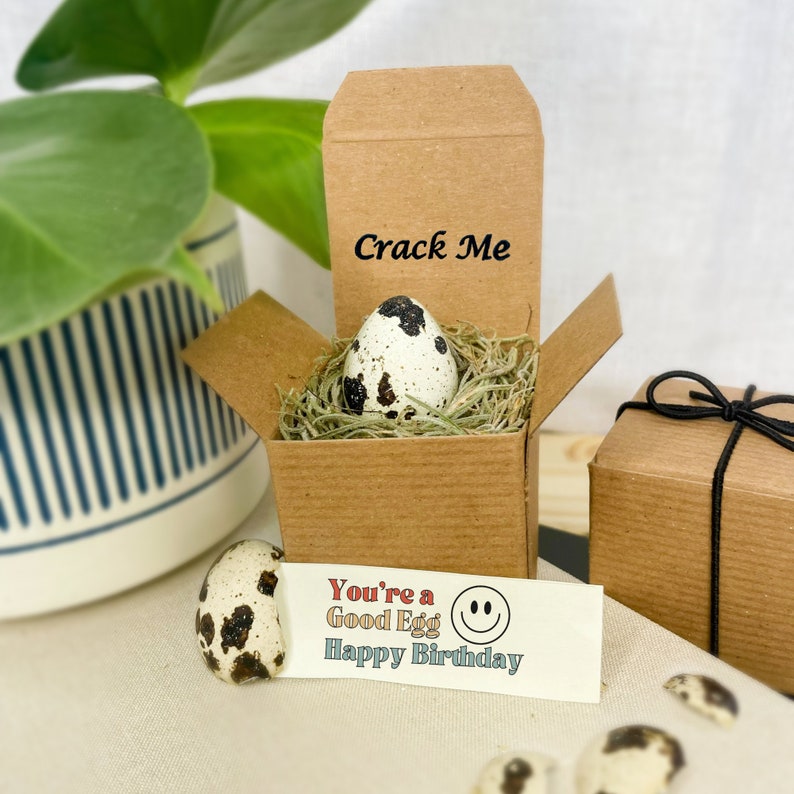 May include: A small brown box with a quail egg inside. The box has the words "Crack Me" written on the top. A card with the text "You're a Good Egg Happy Birthday" and a smiley face is next to the box.