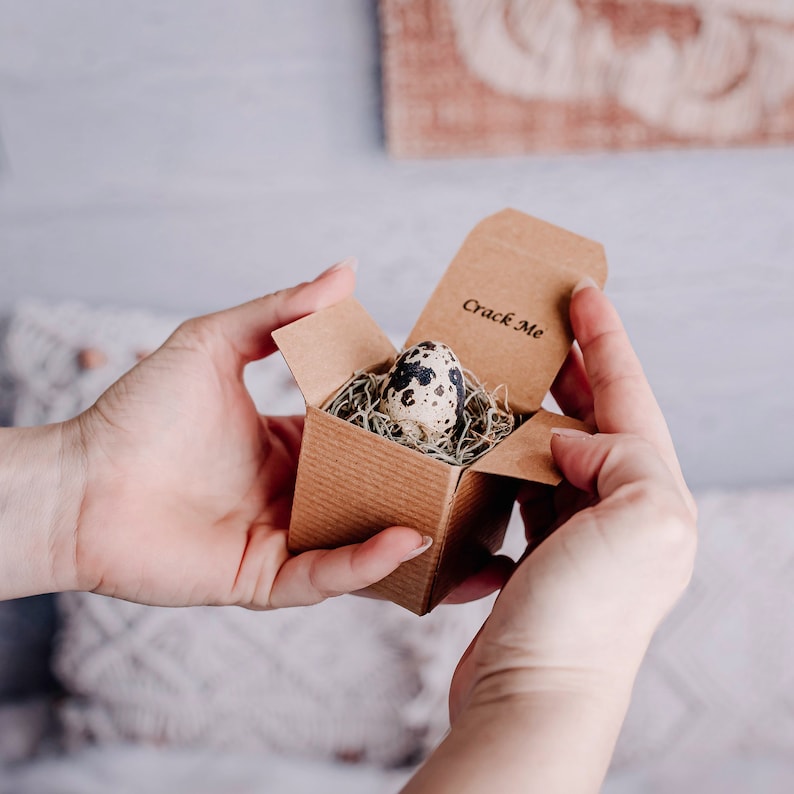 May include: A small brown box with the words "Crack Me" on the lid. The box is open and a speckled quail egg is inside, nestled in a bed of moss.