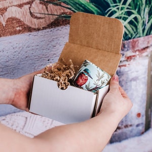 May include: A white gift box with a brown cardboard lid, holding a cylindrical container with a bird illustration. The container is light blue with a red and blue bird design and green foliage. The box contains brown paper filler.