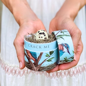 May include: A round, light blue box with a bird and branch design, holding a speckled egg nestled in straw. The box has the text "CRACK ME" around the side. The lid is open, revealing the egg inside.