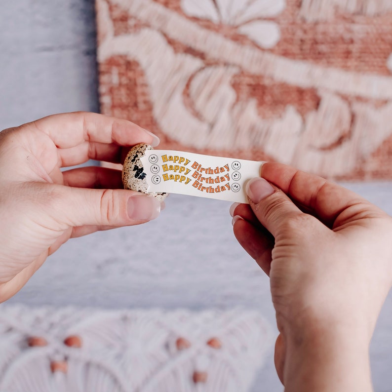 May include: A white paper strip with the text "Happy Birthday" repeated three times, with a small smiley face icon next to each phrase. The strip is being peeled off of a small, speckled, brown quail egg.