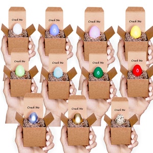 May include: Twelve small cardboard boxes with lids open, each containing a colorful Easter egg. The boxes are labeled "Crack Me" and are arranged in three rows of four boxes.