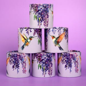 K&ouml;nnte beinhalten: Sechs zylindrische Schachteln, die pyramidenf&ouml;rmig gestapelt sind. Jede Schachtel ist helllila mit einem Blumendesign aus lila Blauregen und einem Kolibri. Der Kolibri hat orange, blaue und gr&uuml;ne Federn. Der Hintergrund ist einfarbig lila.