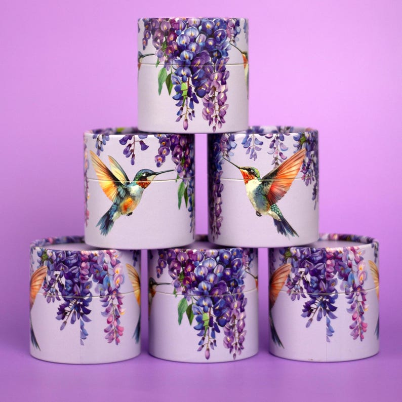K&ouml;nnte beinhalten: Sechs zylindrische Schachteln, die pyramidenf&ouml;rmig gestapelt sind. Jede Schachtel ist helllila mit einem Blumendesign aus lila Blauregen und einem Kolibri. Der Kolibri hat orange, blaue und gr&uuml;ne Federn. Der Hintergrund ist einfarbig lila.