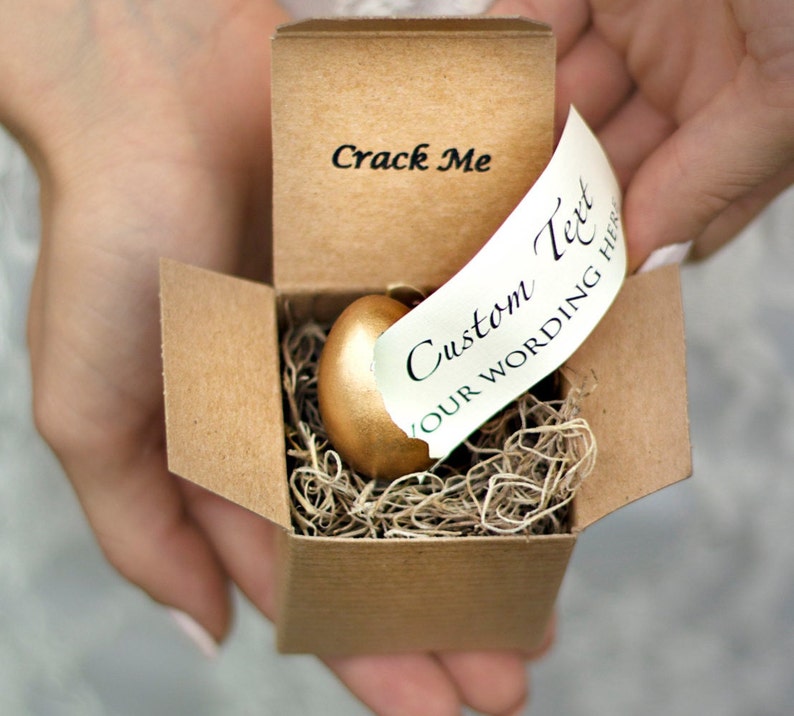 May include: A small cardboard box with a gold egg inside. The box is open and the egg is sitting on a bed of straw. The box has the words "Crack Me" printed on the front. A white tag with the text "Custom Text Your Wording Here" is attached to the box.