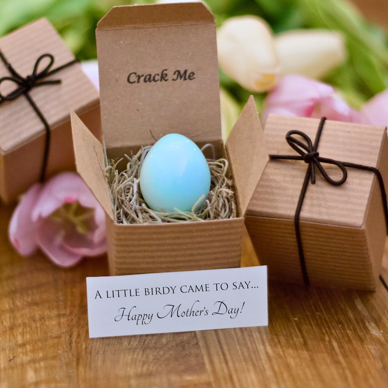 May include: A light blue egg nestled in a cardboard box with the text "Crack Me." The box is filled with straw and is surrounded by two other gift boxes tied with black string. A card reads "A LITTLE BIRDY CAME TO SAY... Happy Mother's Day!"