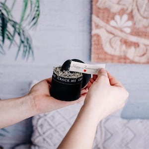 May include: A black round gift box with the text "CRACK ME" on the side. A small paper banner with "Happy Anniversary!" and a red heart is being held up. The box is filled with shredded paper.