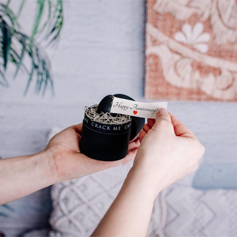 May include: A black round gift box with the text "CRACK ME" on the side. A small paper banner with "Happy Anniversary!" and a red heart is being held up. The box is filled with shredded paper.