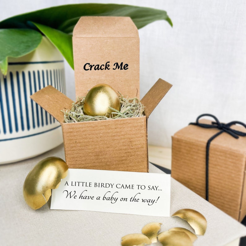 May include: A gold-painted egg in a brown cardboard box with the words "Crack Me" on the lid. The box is open and the egg is surrounded by brown shredded paper. A white card with the text "A little birdie came to say... We have a baby on the way!" is on a white surface next to the box. There are pieces of broken gold-painted egg on the surface.