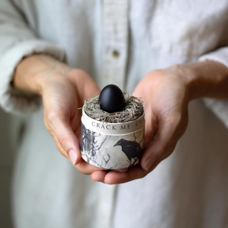 May include: A black egg rests in a small nest inside a cylindrical container. The container is decorated with a raven illustration and the text "CRACK ME". The egg is surrounded by dried grass and held in cupped hands.