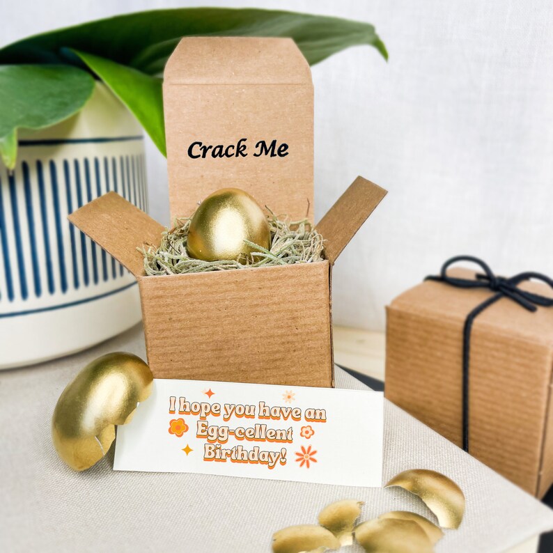 May include: A gold-painted egg in a brown cardboard box with the words "Crack Me" on the lid. The box is surrounded by shredded paper. A card with the text "I hope you have an Egg-cellent Birthday!" is next to the box. The egg is broken into pieces.