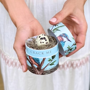 May include: A small, round, light blue gift box with a bird and branch design, holding a speckled egg nestled in dried grass. The box reads "CRACK ME" in white text. The lid is open, revealing the egg.