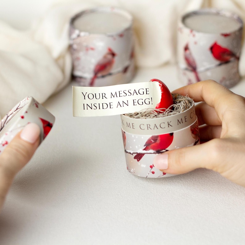 Puede incluir: Peque&ntilde;as cajas de regalo cil&iacute;ndricas con un dise&ntilde;o de p&aacute;jaro cardenal de invierno. Una caja est&aacute; abierta, revelando un huevo rojo y un mensaje que dice "YOUR MESSAGE INSIDE AN EGG!". Las otras cajas est&aacute;n cerradas. Las cajas tienen unos 5 cm de di&aacute;metro.