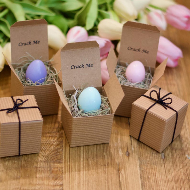 May include: Several small, corrugated cardboard boxes with the text "Crack Me" printed on the inside flaps. Each box contains a pastel-colored egg nestled in straw. Two smaller gift boxes with black string are also present.