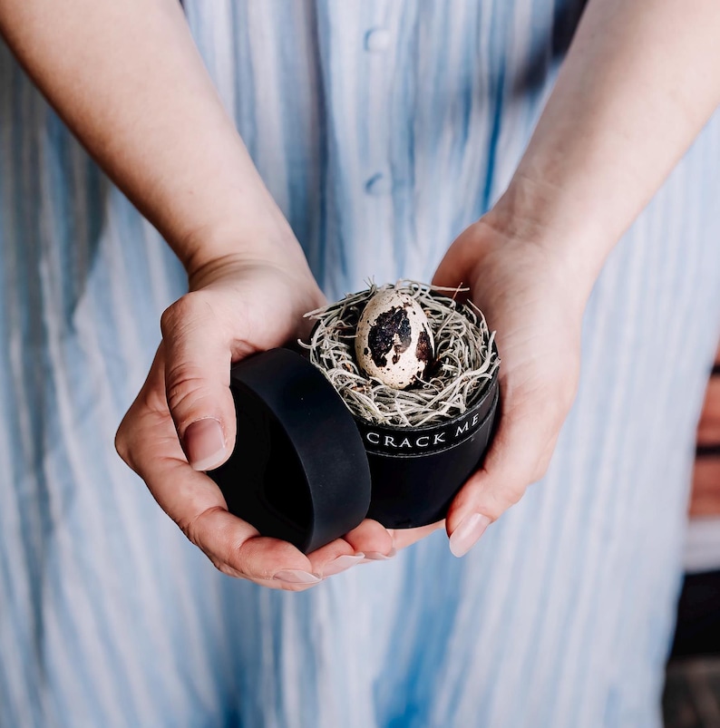 May include: A black box with the text "CRACK ME" on the side, holding a quail egg in a nest of moss.