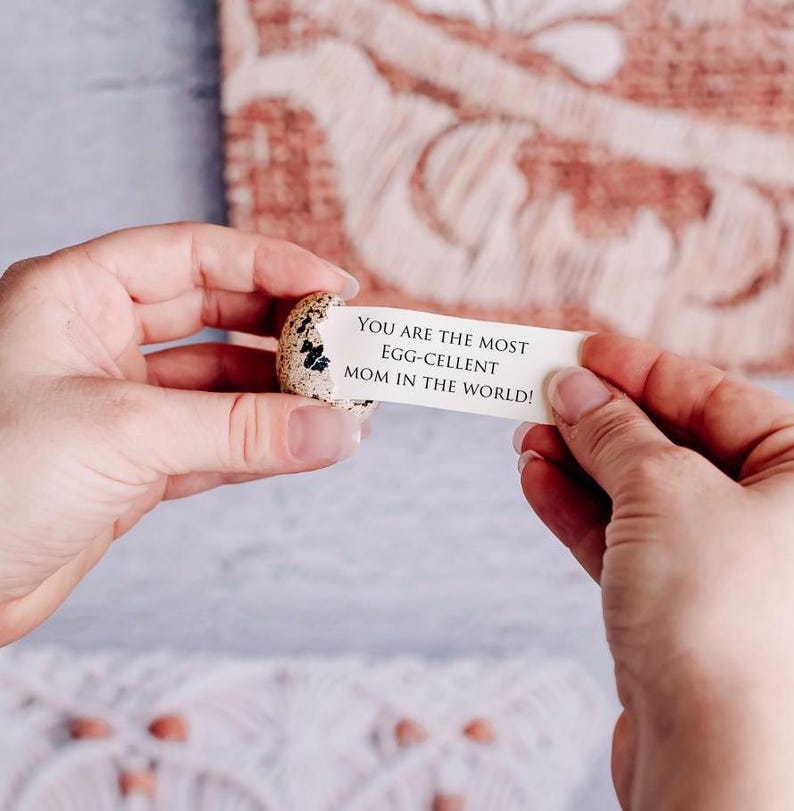 May include: A speckled quail egg with a message reading "YOU ARE THE MOST EGG-CELLENT MOM IN THE WORLD!" held between two hands. The background features a patterned, reddish-brown fabric.