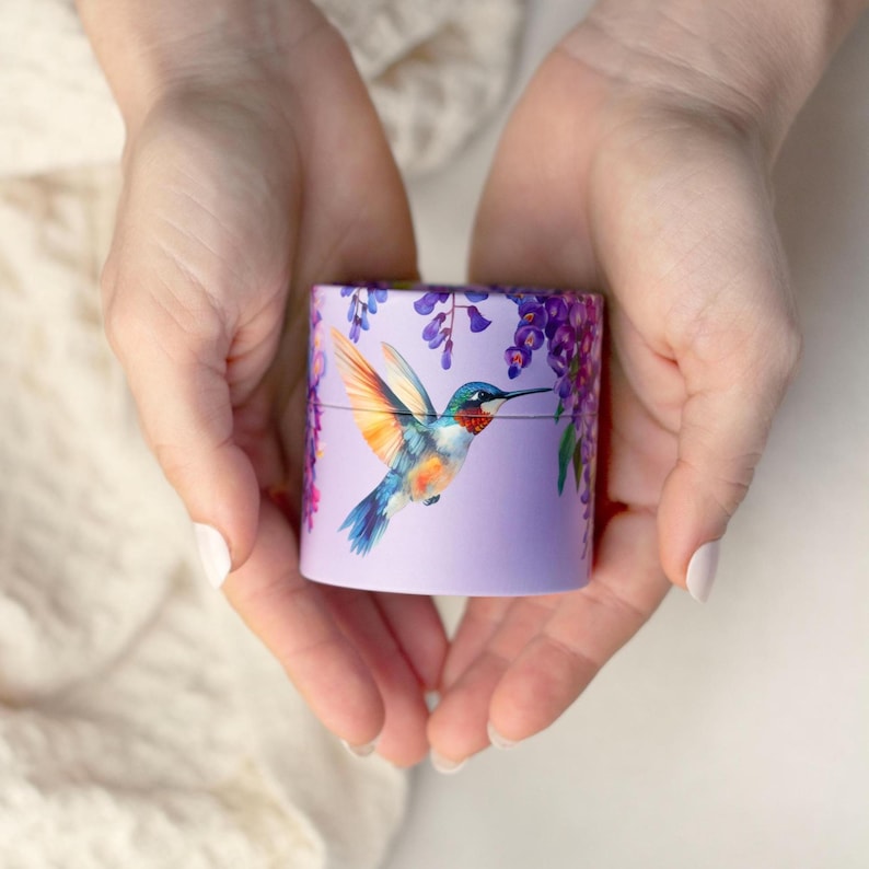 K&ouml;nnte beinhalten: Ein kleiner, zylindrischer Beh&auml;lter mit einem Kolibri- und Blauregen-Design. Der Kolibri hat blaue, orange und wei&szlig;e Federn. Der Beh&auml;lter wird in zwei H&auml;nden gehalten.