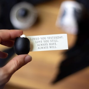 May include: A black, oval-shaped object with a white top is held near a white card with black text. The card reads: "LOVED YOU YESTERDAY, LOVE YOU STILL, ALWAYS HAVE, ALWAYS WILL."