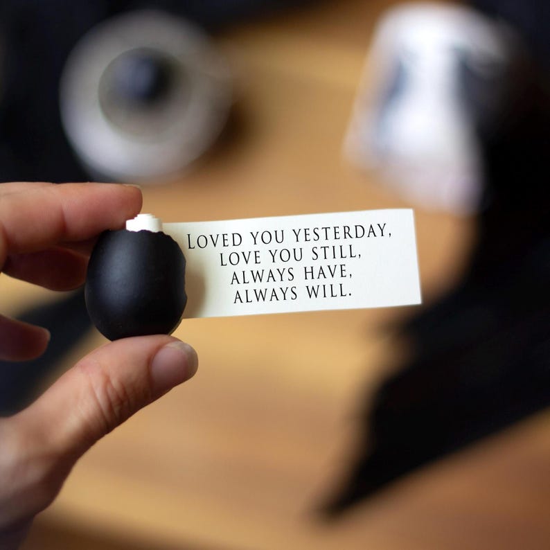 May include: A black, oval-shaped object with a white top is held near a white card with black text. The card reads: "LOVED YOU YESTERDAY, LOVE YOU STILL, ALWAYS HAVE, ALWAYS WILL."