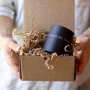 May include: A black cylindrical container with the text "CRACK ME" is nestled in shredded brown paper inside a cardboard box. The box is open, and the container is the focal point.